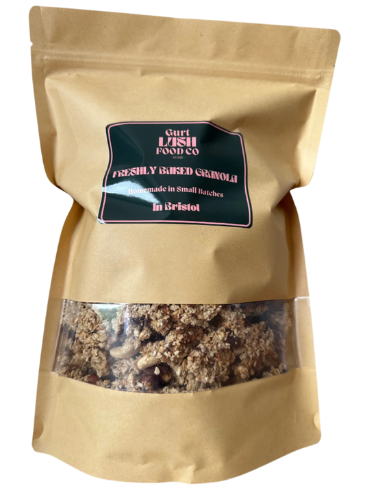 A paper bag of freshly baked granola by Gurt Lush Food Co with a clear window showing the granola inside, labeled as 'The OG' with nuts, seeds, and sultanas.
