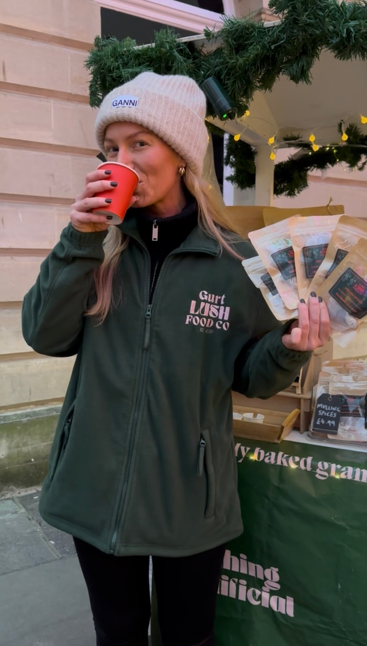 Person wearing a green jacket with visible branding, drinking from a red cup, and holding printed materials.