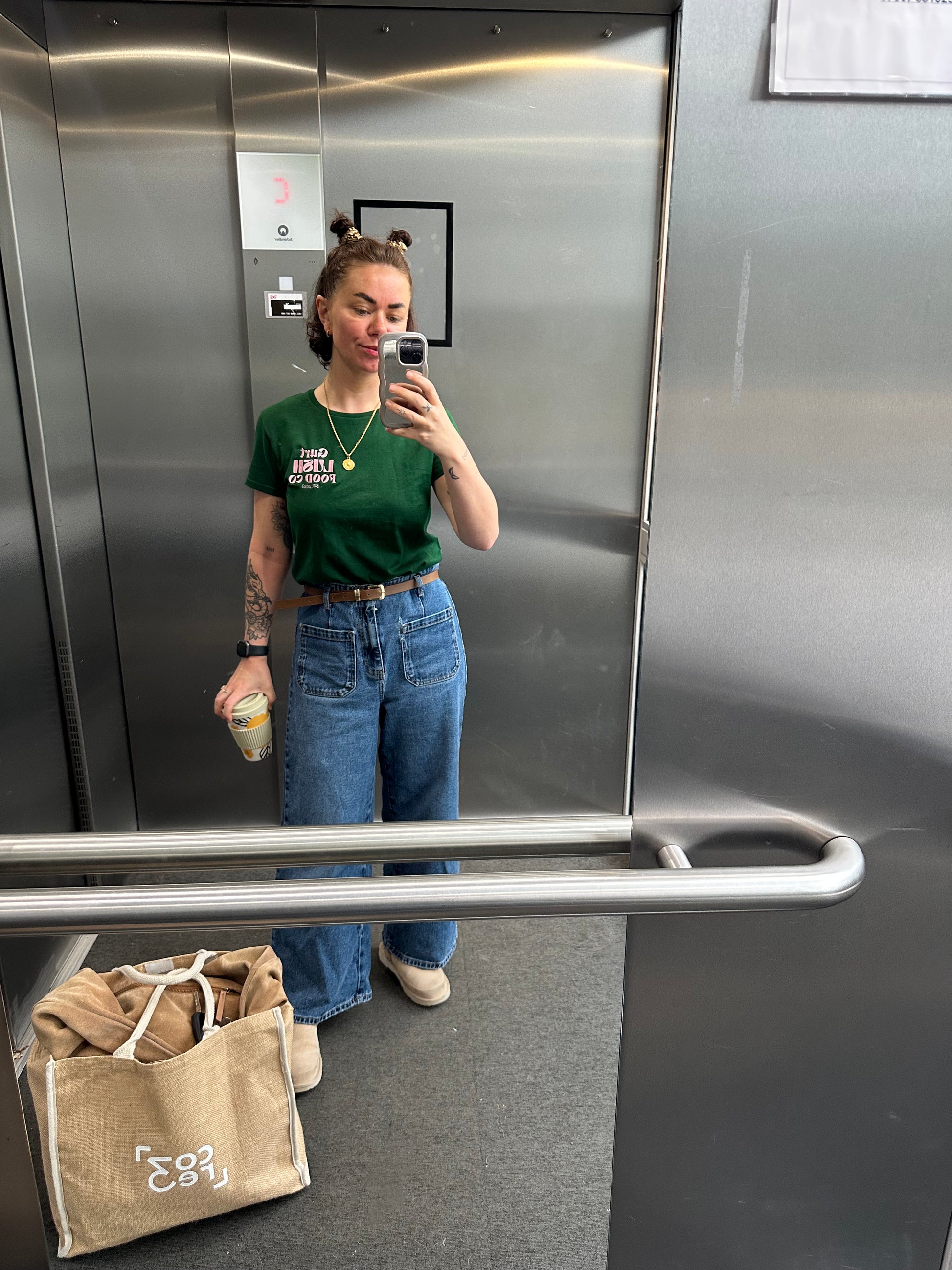 Person taking a photo of themselves in an elevator mirror, wearing a green shirt and blue jeans.